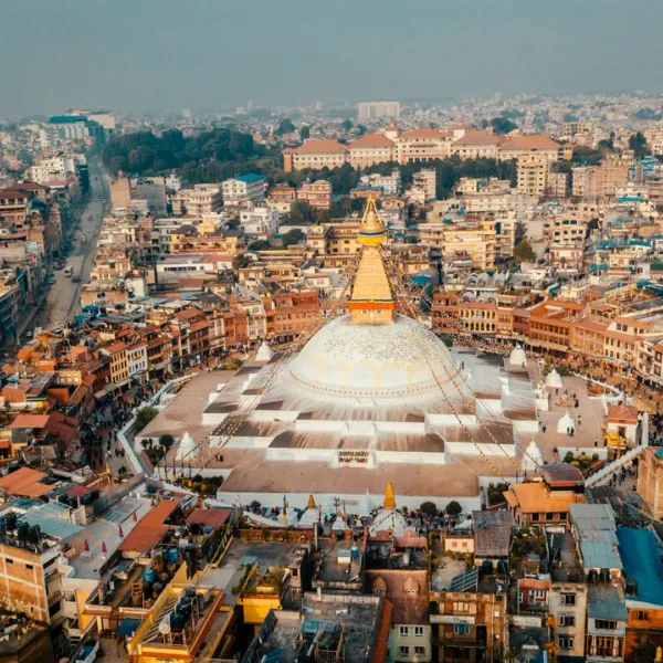 An aerial view of the massive white dome and golden spire of the Boudhanath Stupa in Kathmandu, surrounded by the densely packed buildings of the city.