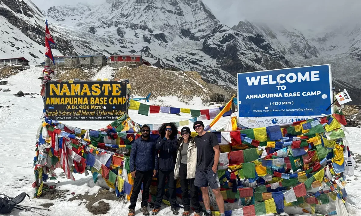 Four hikers at Annapurna Base Camp with prayer flags and snowy mountains.