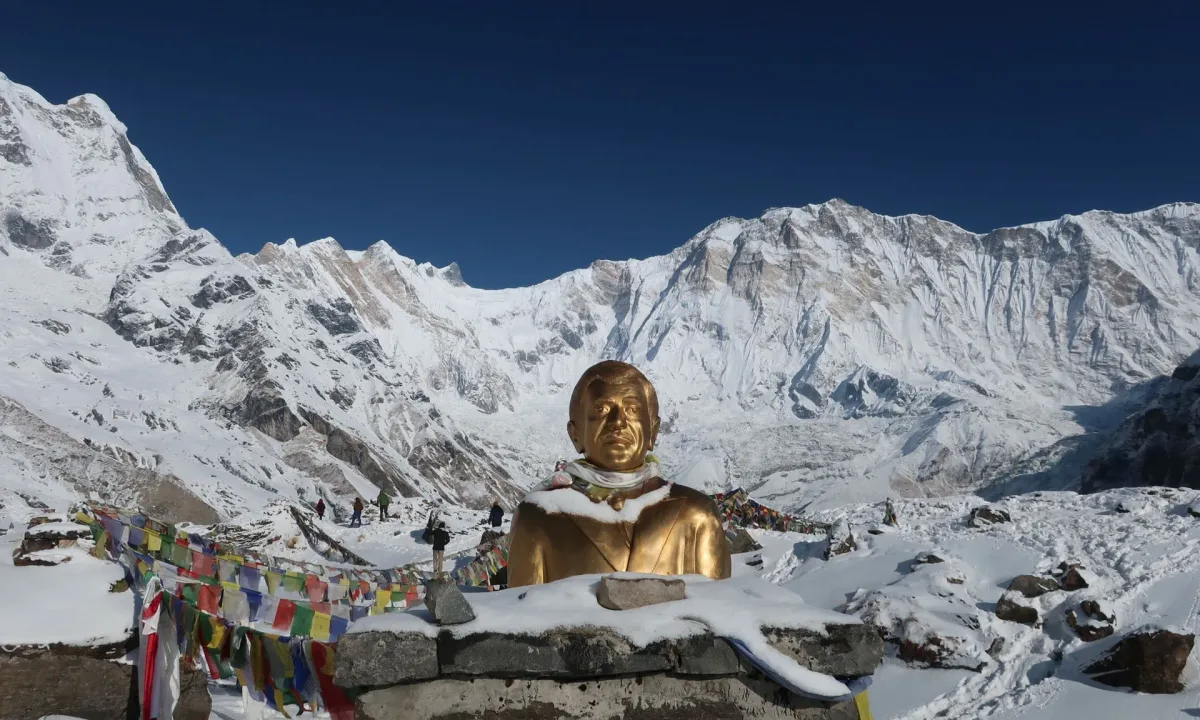 Golden statue at Annapurna Base Camp, Nepal, with snow-capped mountains and prayer flags.