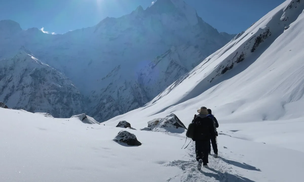 Hikers trek through a snowy mountain landscape with a bright sun and majestic peaks.
