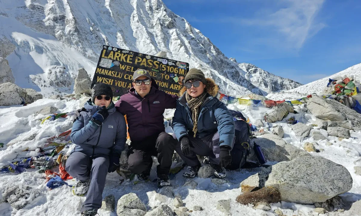 Three people pose at Larkya Pass (5106m) in front of a sign, surrounded by snow and prayer flags.