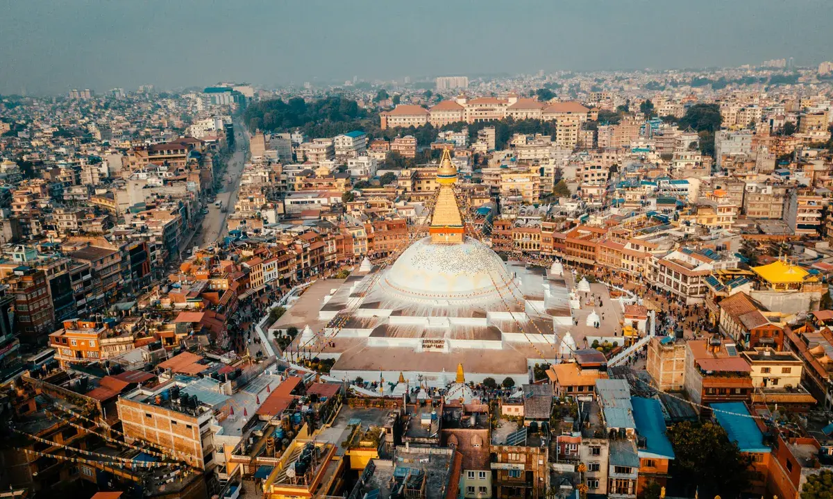 An aerial view of the massive white dome and golden spire of the Boudhanath Stupa in Kathmandu, surrounded by the densely packed buildings of the city.