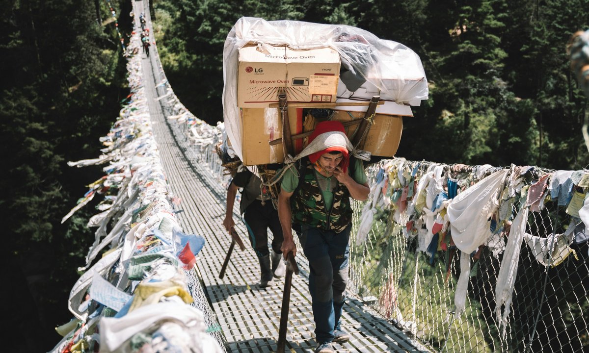 Sherpa porter with heavy load on suspension bridge, prayer flags  Sherpa porter carrying heavy load across suspension bridge with prayer flags EBC Trek Khumbu Nepal