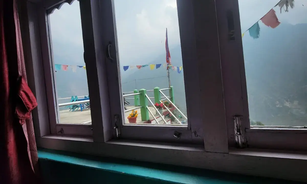 View through a teahouse window in Ulleri showing colourful prayer flags, terrace railing with potted flowers, and misty Himalayan mountains