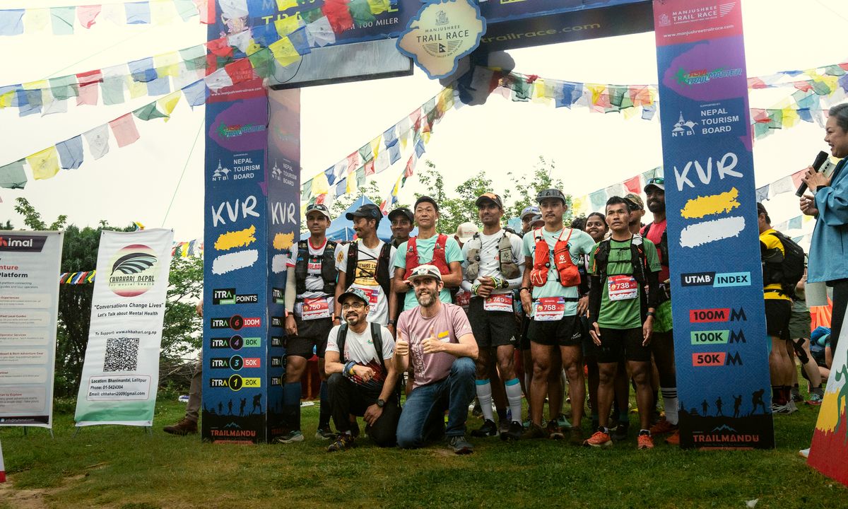 Runners posing at the Manjushree Trail Race start arch with KVR route banners, Nepal Tourism Board sponsorship boards, ITRA points markers, and colourful prayer flags strung overhead