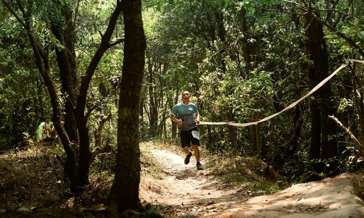 Trail runner with race bib running through a sunlit forest path during a trail race in Nepal, surrounded by tall trees and dappled light
