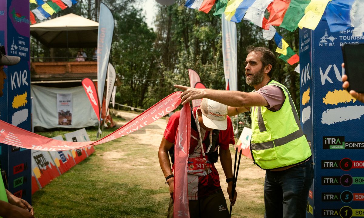 Runner breaking the finish tape at the Manjushree Trail Race KVR route finish line as a race official in a high-vis vest directs, with Nepal Tourism Board banners and prayer flags in the background