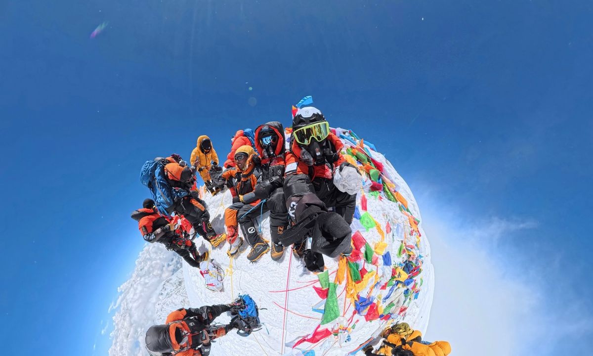 Climbers at the summit of Mount Everest surrounded by colorful prayer flags, captured in a 360-degree fisheye shot during the May 2026 expedition