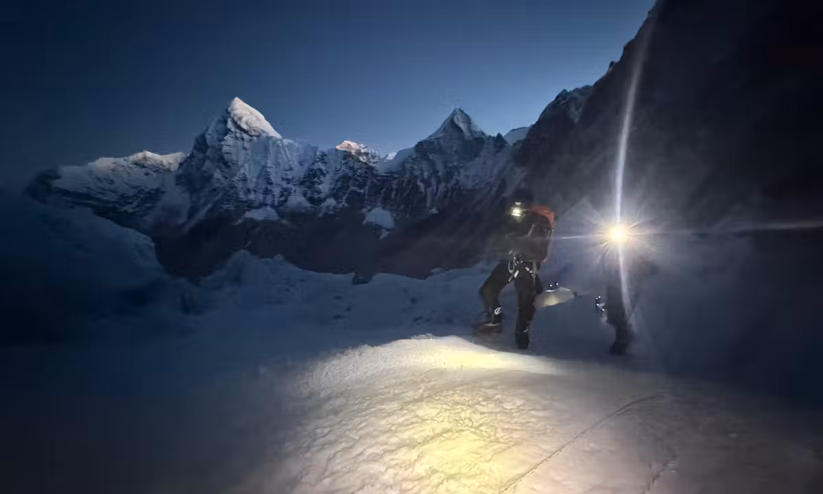 A climber ascending Everest before dawn by headlamp, with the Himalayan peaks rising against a deep blue pre-sunrise sky
