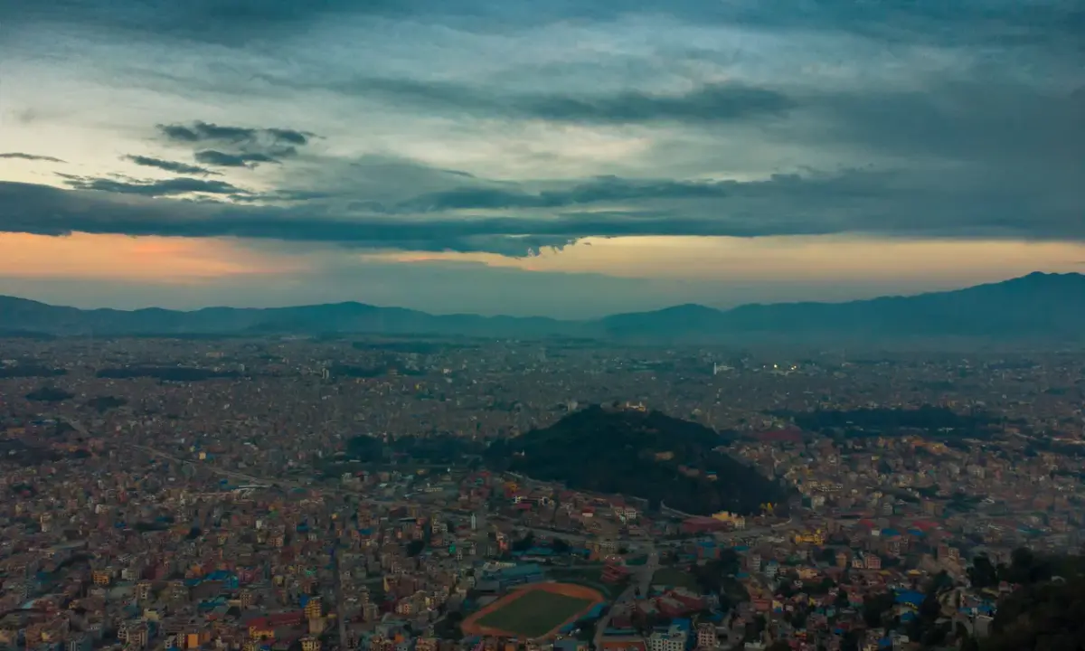 Panoramic aerial view of Kathmandu Valley at dusk with city lights, Swayambhunath hill, and dramatic cloudy sky over distant 