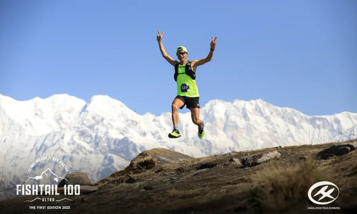 Trail runner jumping joyfully with peace signs against snow-capped Annapurna mountain range during Fishtail 100 Ultra trail r