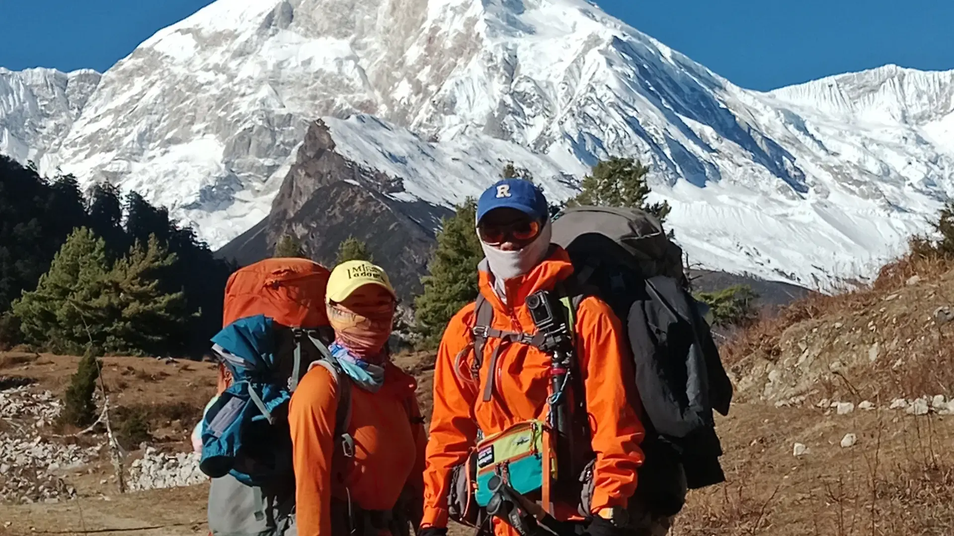 Hikers with large backpacks stand before a majestic snow-capped mountain under a clear blue sky.