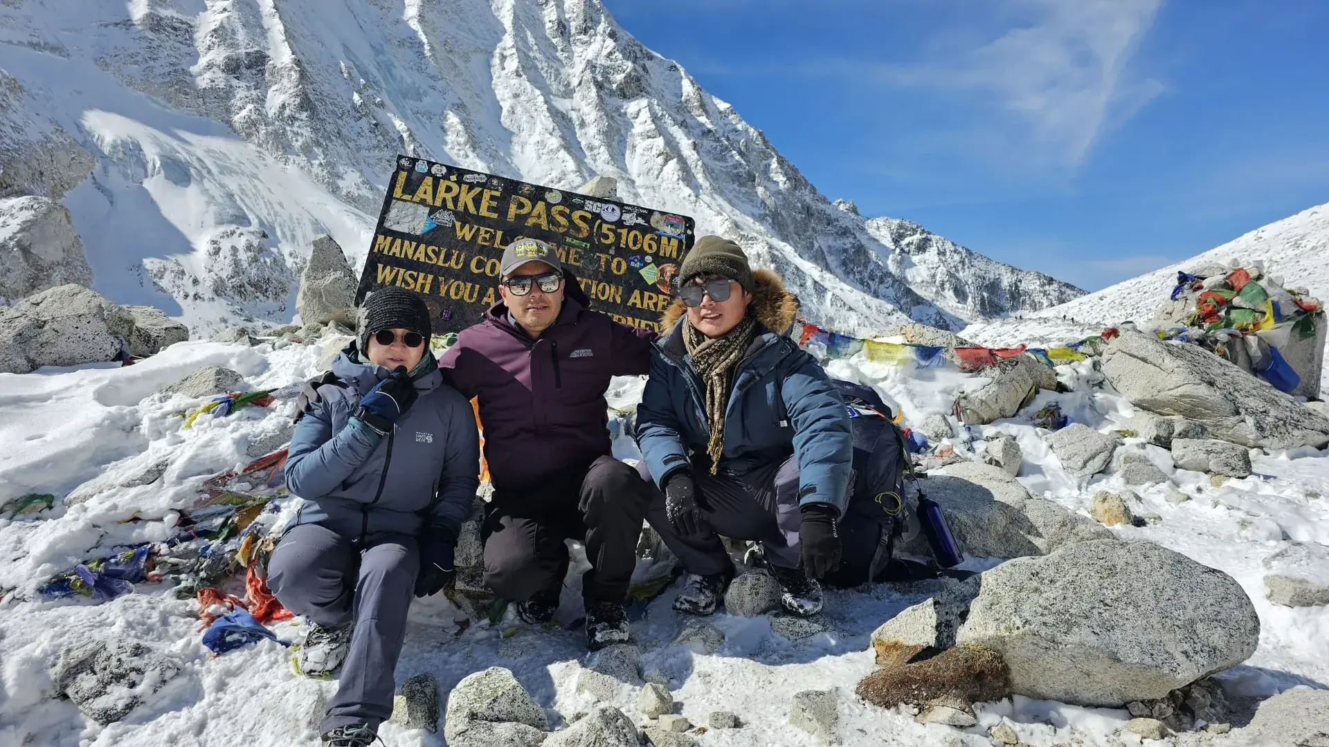 Three people pose at Larkya Pass (5106m) in front of a sign, surrounded by snow and prayer flags.