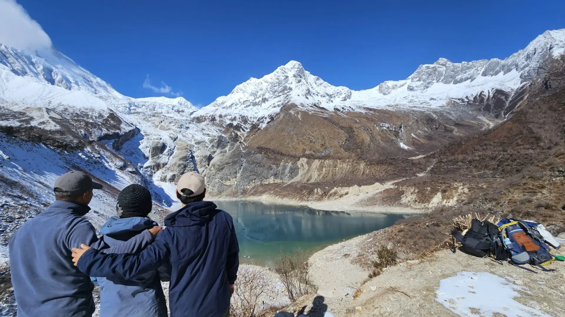 Three hikers admire a serene, turquoise lake surrounded by snow-capped mountains.