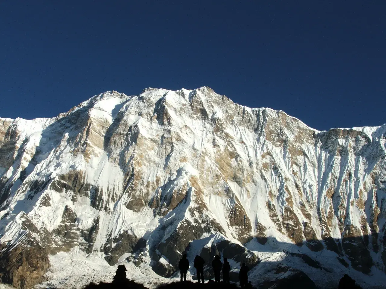 Hikers silhouetted against a massive snow-covered mountain under a clear blue sky.