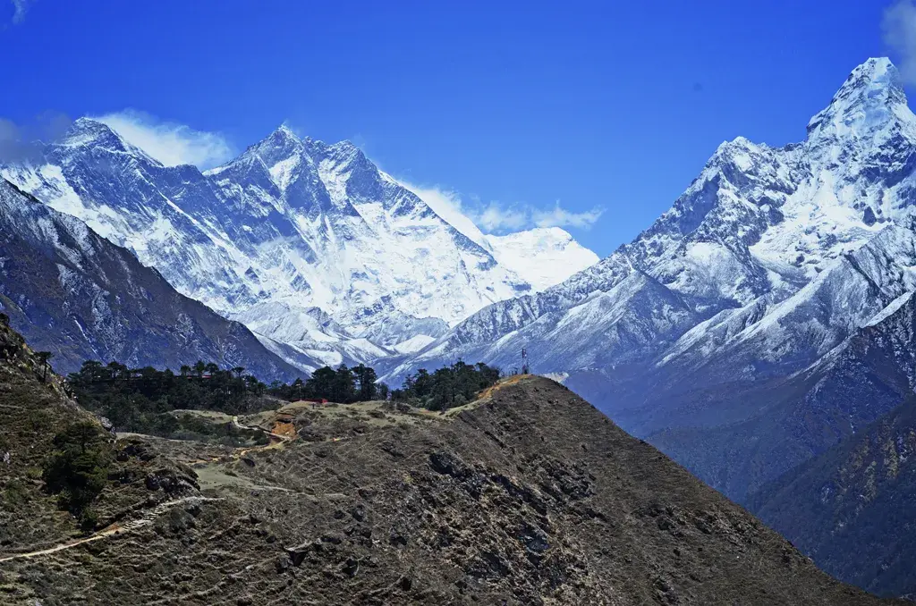 Snow-capped Himalayas loom over a rugged mountain trail and village.