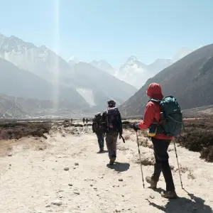 Hikers trek through a dusty mountain valley with snow-capped peaks in the background.
