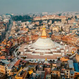 An aerial view of the massive white dome and golden spire of the Boudhanath Stupa in Kathmandu, surrounded by the densely packed buildings of the city.