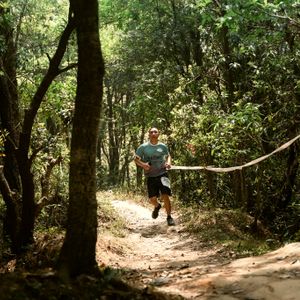 Trail runner with race bib running through a sunlit forest path during a trail race in Nepal, surrounded by tall trees and dappled light