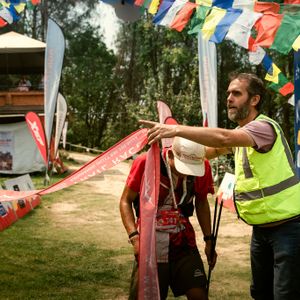 Runner breaking the finish tape at the Manjushree Trail Race KVR route finish line as a race official in a high-vis vest directs, with Nepal Tourism Board banners and prayer flags in the background