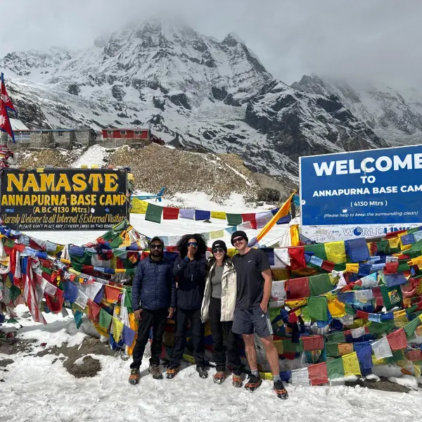 Four hikers at Annapurna Base Camp with prayer flags and snowy mountains.
