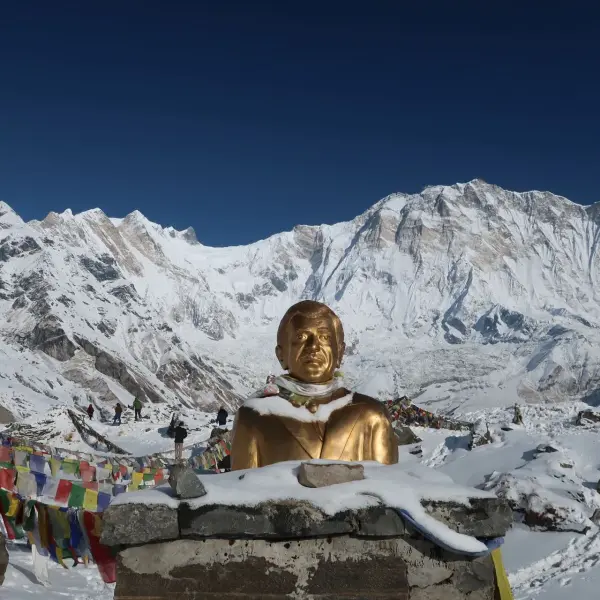Golden statue at Annapurna Base Camp, Nepal, with snow-capped mountains and prayer flags.