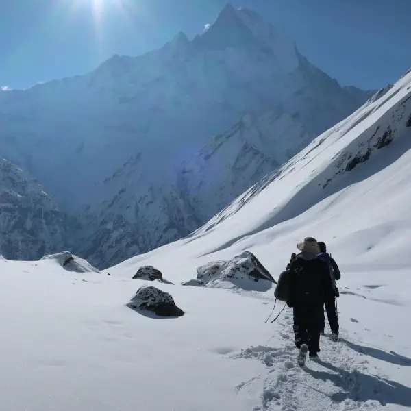 Hikers trek through a snowy mountain landscape with a bright sun and majestic peaks.