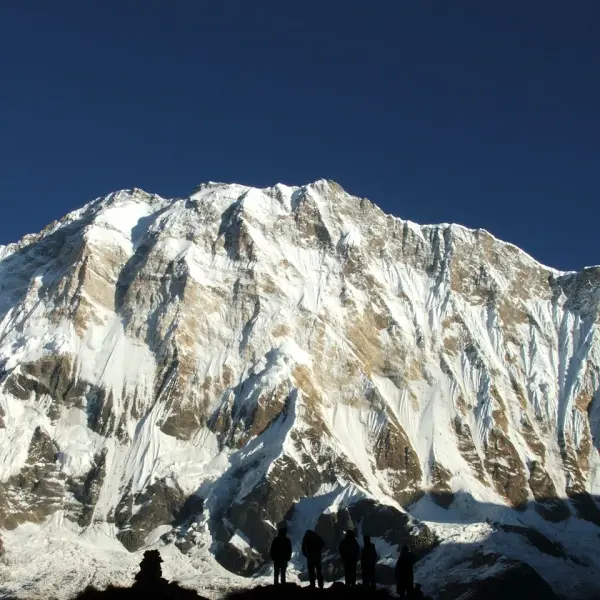 Hikers silhouetted against a massive snow-capped mountain under a deep blue sky.