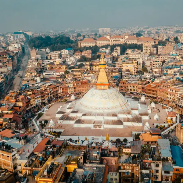 An aerial view of the massive white dome and golden spire of the Boudhanath Stupa in Kathmandu, surrounded by the densely packed buildings of the city.